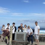 展望台での研究室メンバーの集合写真。中心には展望台の石碑があり、右から先生、栗原さん、新部さん、西田さん、中央の石碑の裏から上半身を出すように佐藤くんが、その左に千野くん、平野くん、柏原さんが並んでいる。