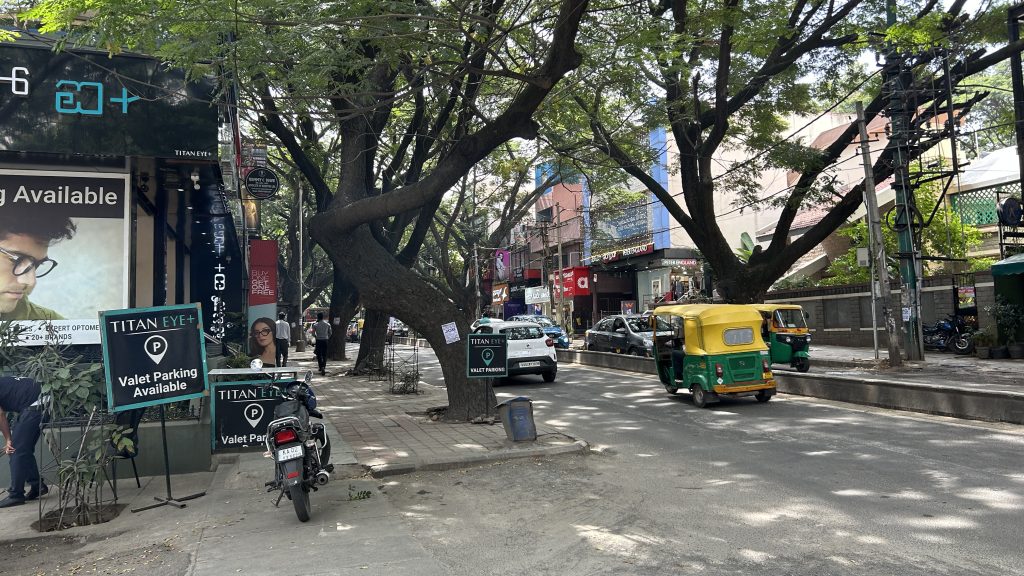 バンガロールの大通りの写真。右下から左奥に向かって大通りが伸びている。手前の道路にはトゥクトゥクや自動車が通行している。道路脇には比較的綺麗な概観の店が並んでいる。