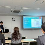 市川が学会発表をしている写真。左奥に市川が登壇しており、その横にスライドを投影したスクリーンが映っている。手前には聴衆が映っている。